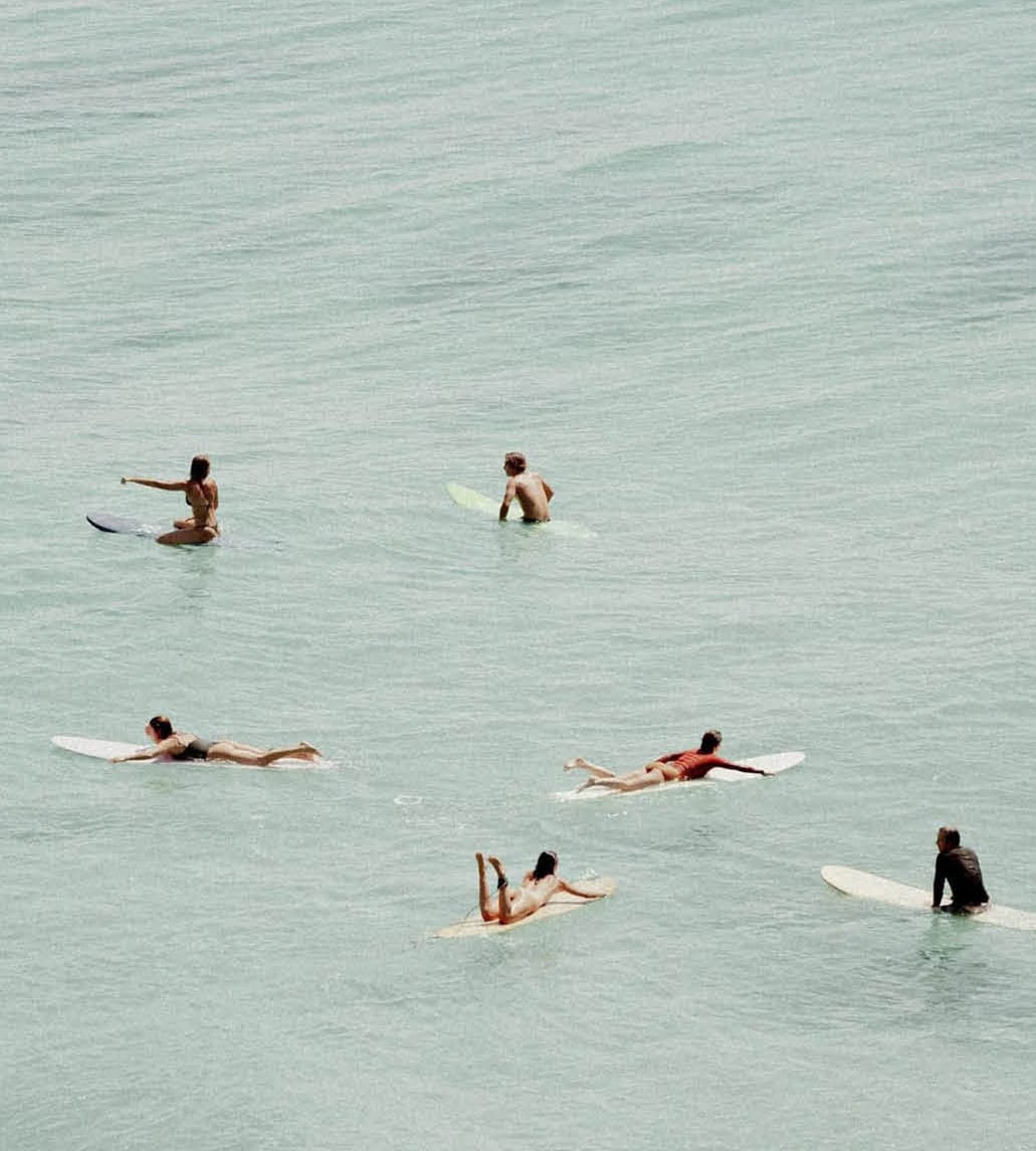 An aerial or high-angle view of six surfers paddling or resting on their longboards in a calm, pale turquoise-green ocean.