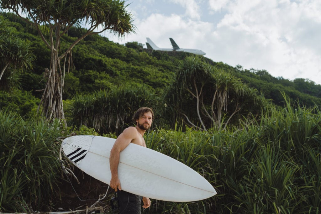 A man carrying a white surfboard stands in front of a lush, tropical hillside.