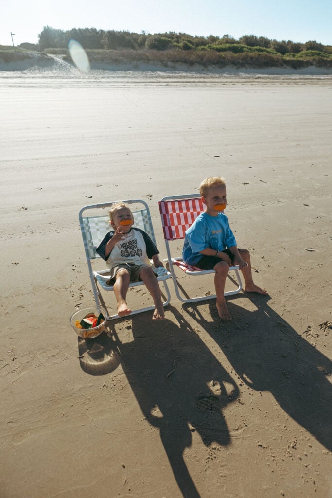 Two young children sitting in beach chairs on a wide, sandy beach
