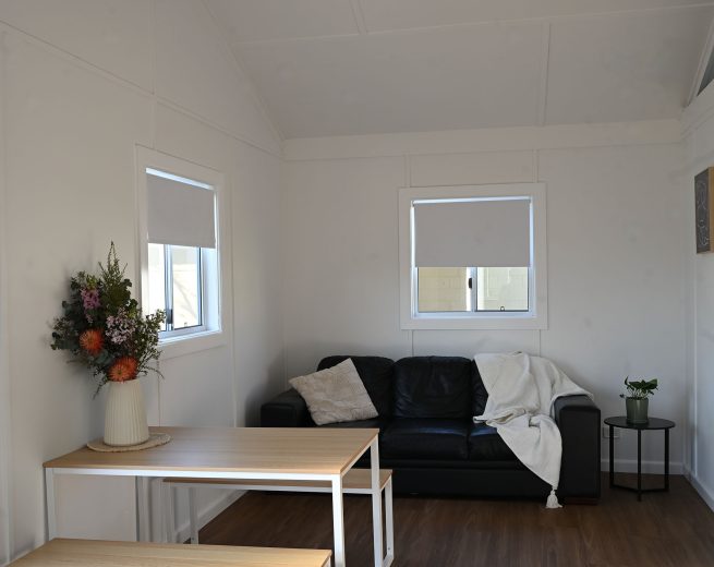 A dark, black leather-look sofa is placed against the wall beneath a window with a light grey roller blind.