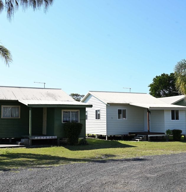 A group of small, simple holiday cabins or bungalows set on a grassy area under a bright, clear blue sky.