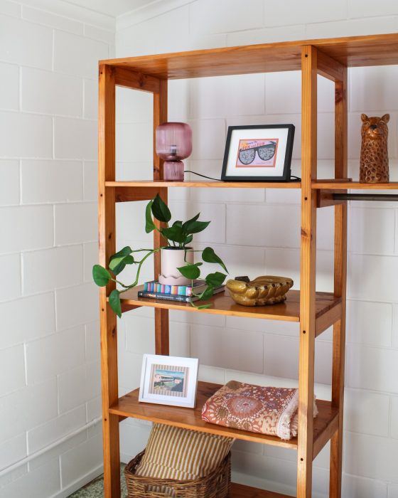 A tall wooden shelving unit against a white brick wall, decorated with a mauve glass lamp, framed art, a pothos plant, a folded patterned textile, and a wicker basket.