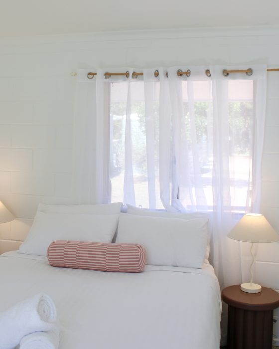 A serene, white-walled bedroom featuring a bed with crisp white linens and a pink striped bolster pillow.