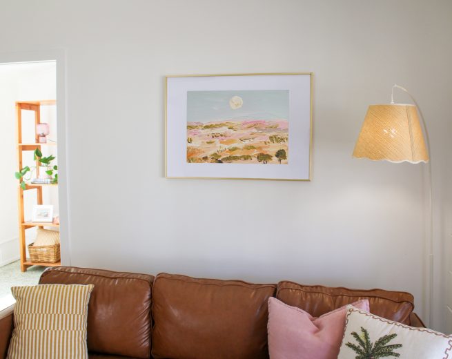 A brown leather sofa with decorative pillows sits under a framed landscape painting and a scalloped floor lamp