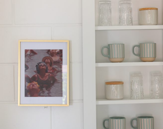 A framed photo of people swimming in a pool hangs on a white brick wall next to white built-in shelves containing striped mugs and crystal glassware.