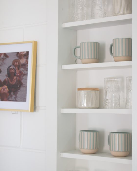 Striped mugs, crystal glasses, and ceramic jars arranged on white built-in shelves next to a framed photo on a white brick wall.