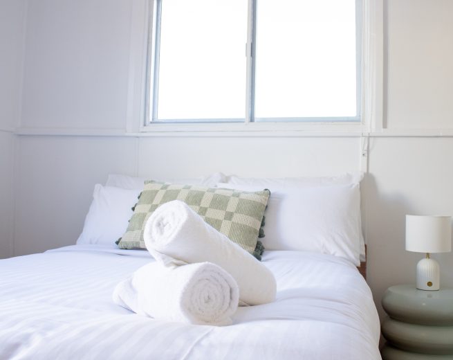 A white bed with rolled towels and a green chequered pillow, positioned next to a pale blue sculptural nightstand under a bright window.