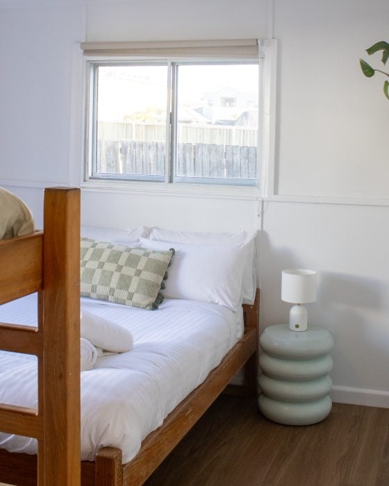 A white bed with rolled towels and a green chequered pillow, positioned next to a pale blue sculptural nightstand under a bright window.