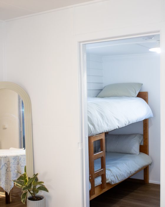 A view through a doorway into a bedroom with wooden bunk beds and light blue linens, with a tall mirror and potted plants in the foreground.