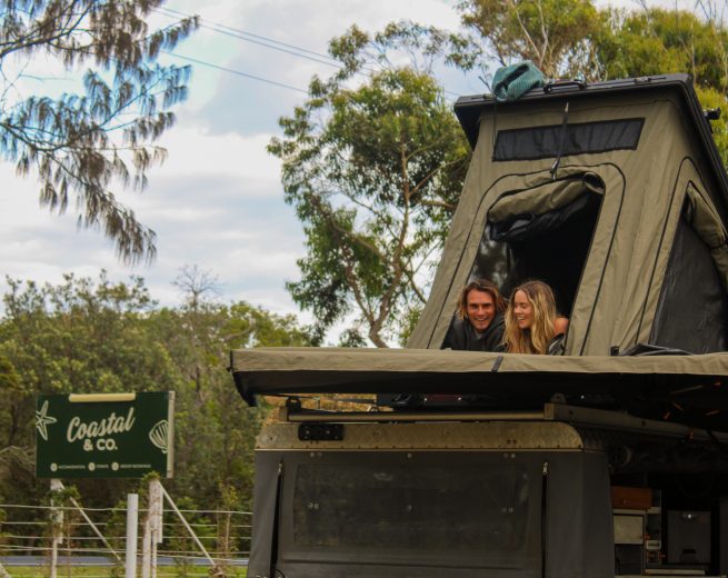 A smiling couple looks out from the opening of an olive green rooftop tent mounted on a vehicle.