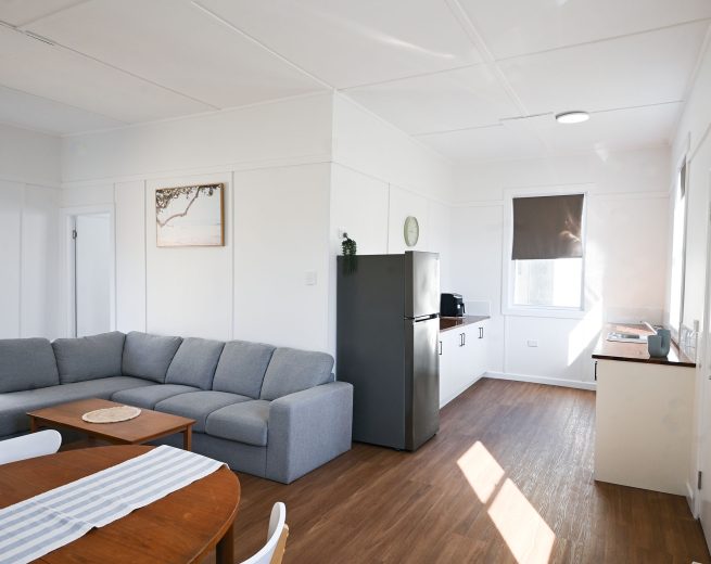 A bright, open-plan living, dining, and kitchen area with white walls and warm wood-look flooring.