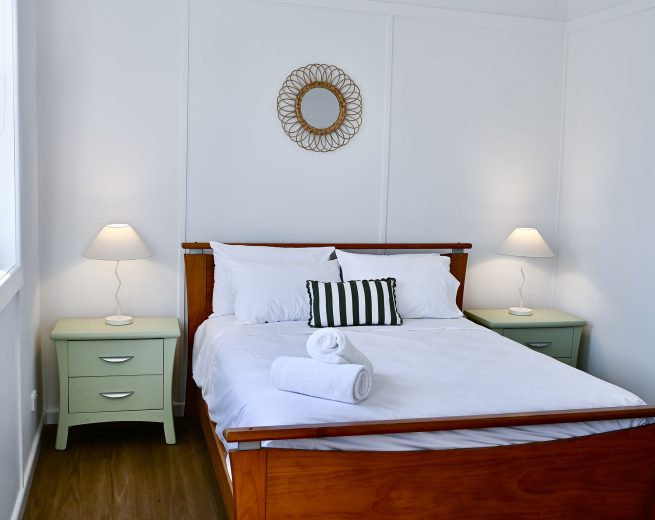 The bed is covered in white linens with a white and black striped bolster pillow and two rolled white towels placed at the foot.