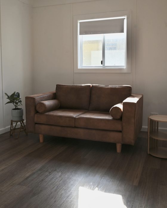 A vertical interior shot of a minimalist living space with a brown leather-look two-seater sofa placed centrally against a white wall.