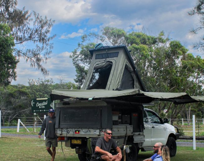 A group of friends relaxing on a picnic blanket and camping chairs next to an off-road truck with a deployed rooftop tent and side awning.