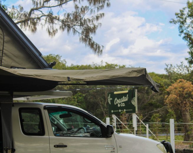 A side view of a white Toyota Hilux truck equipped with an extended tan awning and a rooftop tent, parked in front of a "Coastal & Co." sign.